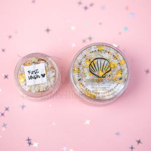 May include: Two clear circular containers filled with iridescent gold and white cosmetic glitter. The smaller container features a white label with the text First Wish and a mouse icon. The larger container lid displays a seashell graphic.