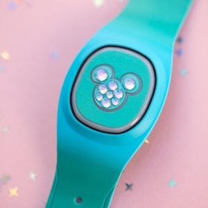 May include: A teal smart wristband with a Mickey Mouse icon made of sparkling gems. The band is on a pink background with star-shaped confetti. The wristband is a wearable tech accessory.