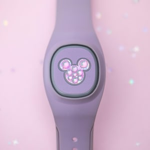 May include: A lavender smart band with a Mickey Mouse icon. The icon is made of iridescent, pearlescent beads. The band is a solid lavender color and has a button and holes for adjustment. The background is a soft pink with glitter.