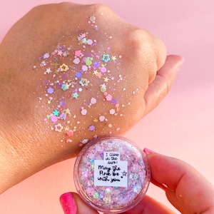 May include: A clear, round container filled with pink and iridescent glitter, with a scattering of glitter on a hand. The container has a label that reads "I glow in the dark! May the Pink be with you."