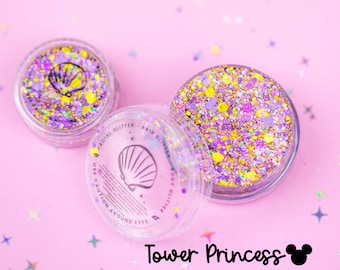 May include: Two clear circular jars filled with purple, gold, and iridescent chunky glitter. The lids feature a black seashell icon and text reading "FACIAL GLITTER HAIR GLITTER BODY GLITTER USE CAUTION AROUND EYES". Text below reads "Tower Princess".