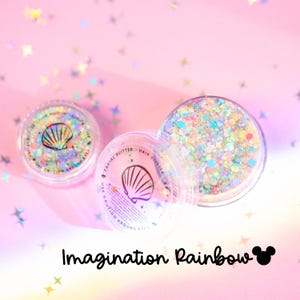 Puede incluir: Dos recipientes circulares transparentes llenos de purpurina iridiscente multicolor para rostro y cabello. Las tapas presentan un gráfico de concha negra y el texto "FACIAL GLITTER - HAIR GLITTER" y "USE CAUTION AROUND EYES". El texto "Imagination Rainbow" aparece debajo.
