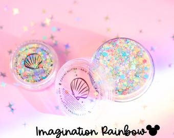 Puede incluir: Dos recipientes circulares transparentes llenos de purpurina iridiscente multicolor para rostro y cabello. Las tapas presentan un gráfico de concha negra y el texto "FACIAL GLITTER - HAIR GLITTER" y "USE CAUTION AROUND EYES". El texto "Imagination Rainbow" aparece debajo.