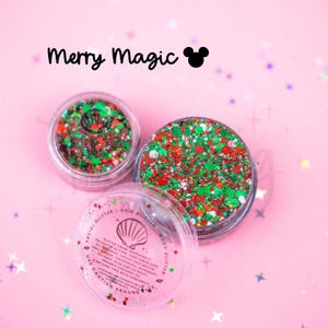 May include: Two clear circular jars filled with red, green, and white glitter. The text Merry Magic with a mouse ear icon appears above. The lid of the open jar features a seashell logo and text listing ingredients and safety warnings.