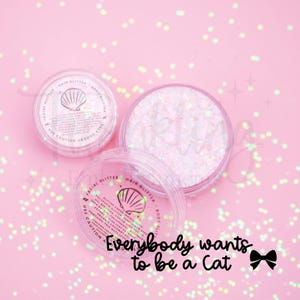 May include: Two clear circular containers filled with iridescent pink glitter balm. Each lid features a seashell icon and text listing ingredients like sunflower seed oil, beeswax, and shea butter. The text reads: Everybody wants to be a Cat.