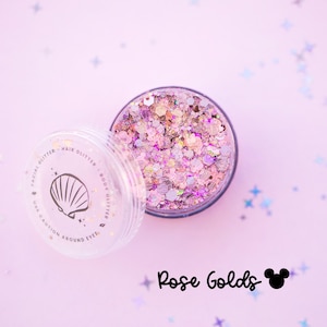 Puede incluir: Un tarro redondo de plástico transparente lleno de purpurina cosmética gruesa en tonos oro rosa, rosa y plata. La tapa tiene un icono de concha y el texto: FACIAL GLITTER HAIR GLITTER BODY GLITTER USE CAUTION AROUND EYES. Debajo dice Rose Golds.