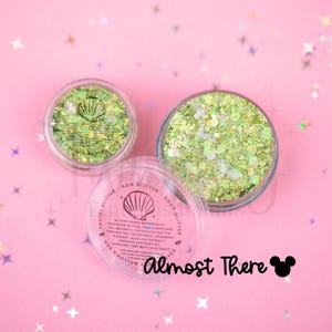 May include: Two clear circular containers filled with lime green and iridescent chunky glitter. The lids feature a seashell icon, the text "Facial Glitter, Hair Glitter, Body Glitter," ingredient lists, and the phrase "Almost There" with a mouse icon.