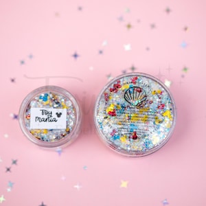 Puede incluir: Dos recipientes circulares transparentes llenos de purpurina gruesa multicolor. El recipiente más pequeño tiene una etiqueta blanca que dice "Toy mania" con un icono de ratón. La tapa del recipiente grande muestra un logo de concha.