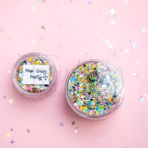 May include: Two clear circular containers filled with multi-colored cosmetic glitter. The smaller container features a white label with the handwritten text Pop Quiz Party ABC. The larger container displays a black shell icon and printed text.