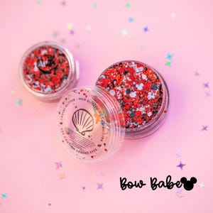 Puede incluir: Dos frascos circulares transparentes llenos de purpurina gruesa roja, negra y blanca. Las tapas presentan un icono de concha, el texto FACIAL GLITTER HAIR GLITTER y USE CAUTION AROUND EYES. Debajo aparece Bow Babe con un icono de orejas.