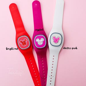 May include: Three colorful wearable wristbands in bright red, crystal, and electric pink. Each band features a circular design with a Mickey Mouse head. The bright red band has the text "bright red", the crystal band has the text "crystal", and the electric pink band has the text "electric pink".