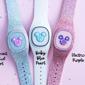 May include: Three sparkly Disney MagicBands with different colored glitter and Mickey Mouse charms. The bands are labeled "Iridescent Orchid Pearl", "Baby Blue Pearl", and "Electric Purple".