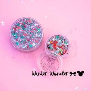 May include: Two clear circular containers filled with multi-colored chunky glitter in shades of red, green, and silver. The lids feature a shell logo and text. The text Winter Wonder is printed below the containers with a bow and mouse icon.