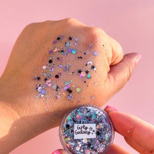 May include: A pot of glitter labelled "Girly Galaxy" containing a mix of iridescent, black, and pastel-coloured glitter. Some glitter is scattered on a hand, against a pink backdrop.