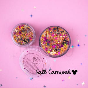 May include: Two clear circular jars filled with multicolored cosmetic glitter in shades of orange, pink, black, and gold, featuring small mouse-ear and seashell shapes. The bottom jar label reads Fall Carnival with a mouse-ear icon.