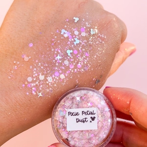 May include: A small clear jar labeled Pixie Petal Dust containing pink and iridescent cosmetic glitter. The glitter features various shapes including small stars, hexagons, and mouse head silhouettes, applied to skin in a shimmering, scattered pattern.