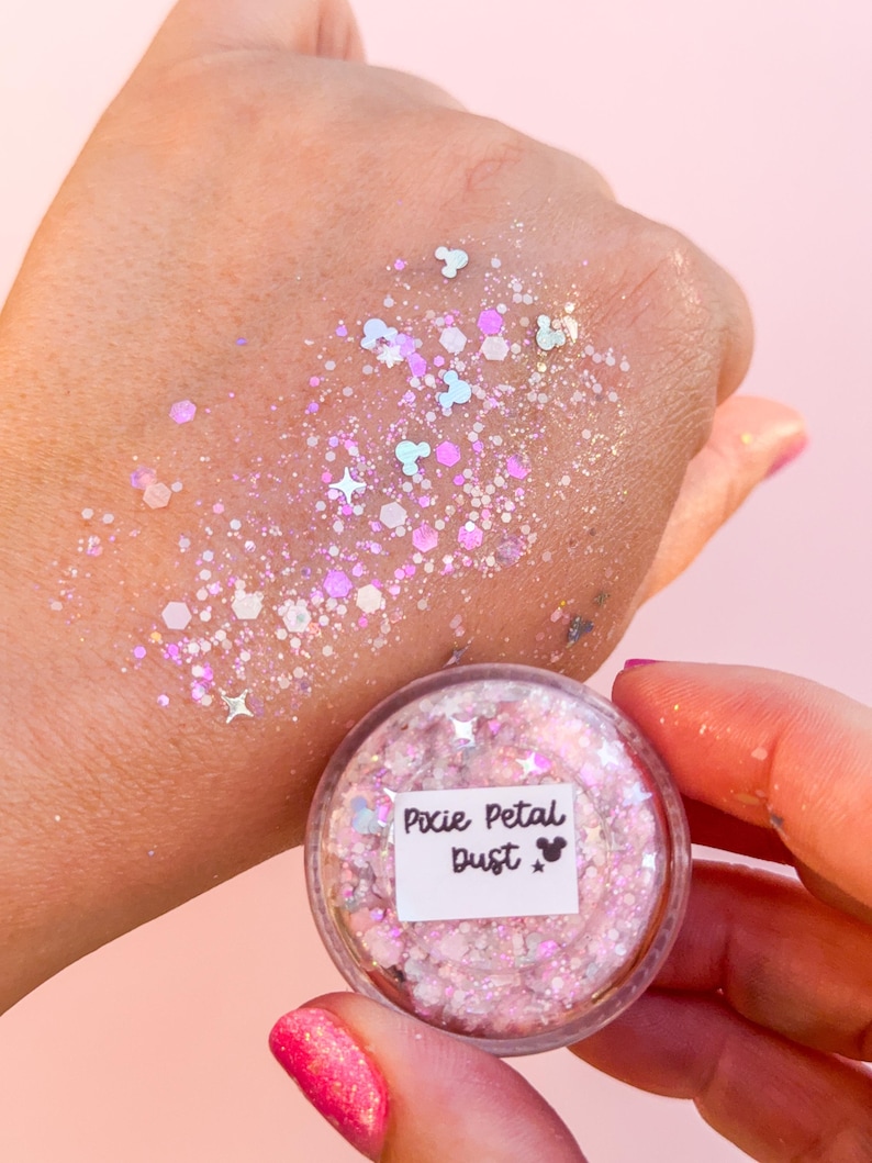 May include: A small clear jar labeled Pixie Petal Dust containing pink and iridescent cosmetic glitter. The glitter features various shapes including small stars, hexagons, and mouse head silhouettes, applied to skin in a shimmering, scattered pattern.