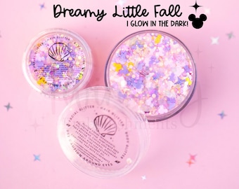 May include: Three clear circular containers filled with multi-colored facial, hair, and body glitter in shades of purple, pink, and yellow. The lids feature a seashell icon and text reading Dreamy Little Fall I GLOW IN THE DARK!