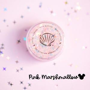 Op de afbeelding: Een ronde doorzichtige pot gevuld met iriserende roze, blauwe en witte glitters. Het deksel bevat een zwarte schelpgrafiek, sterren en de tekst FACIAL GLITTER, HAIR GLITTER, BODY GLITTER en USE CAUTION AROUND EYES. Daaronder staat de tekst Pink Marshmallow.