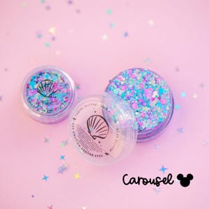 May include: Three clear plastic jars of facial glitter with a seashell design on a pink background. The glitter is a mix of blue, purple, and pink. The text "Carousel" and a Mickey Mouse icon are in the bottom right corner.
