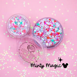 Puede incluir: Dos frascos circulares transparentes llenos de purpurina cosmética multicolor en tonos rosa, blanco y turquesa, con formas de estrellas y conchas. La tapa del frasco abierto muestra texto que incluye "Facial Glitter" y "Body Glitter".