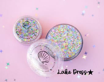 Puede incluir: Tres botes transparentes de purpurina sobre un fondo rosa con confeti en forma de estrella. Los botes contienen purpurina iridiscente y de colores pastel. Un bote tiene un logotipo de concha y el texto "FACIAL GLITTER HAIR GLITTER BODY GLITTER CAUTION AROUND EYES". El texto "Lake Dress" también es visible.