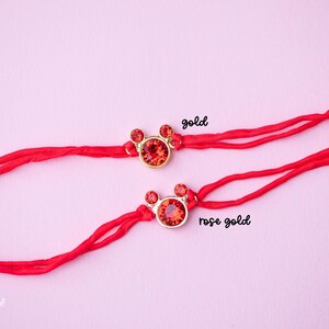 May include: Two red string bracelets with a red crystal charm in the shape of a Mickey Mouse head. The charm on the top bracelet is gold, and the charm on the bottom bracelet is rose gold.