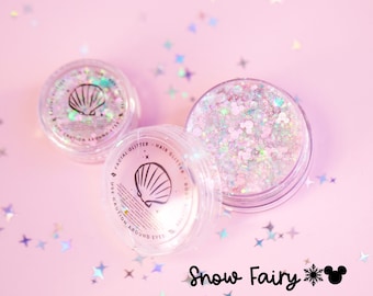 May include: Two clear circular jars filled with iridescent facial and hair glitter. The lids feature a black seashell icon and text reading "Facial Glitter, Hair Glitter, Use Caution Around Eyes." The text "Snow Fairy" appears below.