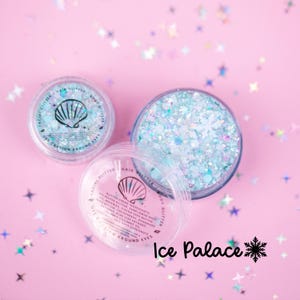 May include: Two clear circular jars containing iridescent light blue and white glitter creme. One jar is open, revealing snowflake-shaped sequins. The lids feature a seashell icon and text reading Facial Glitter, Hair Glitter, Body Glitter.