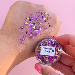 May include: A small clear jar of cosmetic glitter labeled Phoenix's Pride, containing a mix of purple, pink, and gold hexagonal and Mickey Mouse-shaped glitter particles, with a swatch of the same multi-colored glitter applied to the skin.
