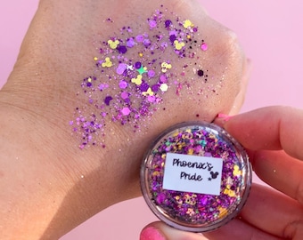 Puede incluir: Un pequeño frasco transparente de purpurina cosmética con la etiqueta Phoenix's Pride, que contiene una mezcla de partículas de purpurina hexagonales y con forma de Mickey Mouse en colores morado, rosa y dorado, con una muestra en la piel.