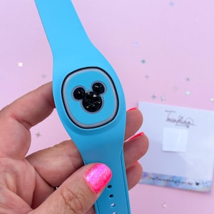 May include: A light blue wearable tech band with a Mickey Mouse design. The band features a square face with a black Mickey Mouse silhouette. The band is held by a person with pink nail polish.