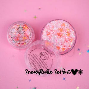 May include: Two round clear plastic jars filled with pink and white iridescent body glitter. The lids feature a seashell icon and printed ingredient text. The text Snowflake Sorbet is written below the jars on a pink background.