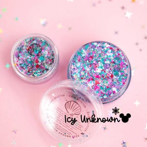 May include: Two clear circular containers filled with multi-colored chunky glitter in shades of teal, pink, purple, and white. The lids feature a black seashell icon and the text Icy Unknown with a mouse ear silhouette icon.