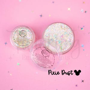 May include: Two clear circular containers filled with iridescent facial and body glitter. Each lid features a seashell icon and text reading: Facial Glitter, Hair Glitter, Body Glitter, Use Caution Around Eyes. Pixie Dust is written below.