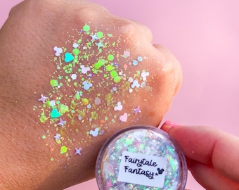 May include: A small clear circular container filled with iridescent cosmetic glitter, featuring a white label with the text Fairytale Fantasy and a mouse head icon. The glitter consists of neon green, pink, and white shapes including stars and hearts.