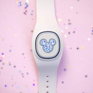 Pode incluir: Pulseira inteligente branca com um design do Mickey Mouse. O design do Mickey Mouse é feito de strass azuis. A pulseira está em um fundo rosa com confetes em forma de estrela.