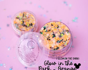May include: Two clear circular jars filled with orange, black, and white chunky glitter. The text on the lid reads: I GLOW IN THE DARK! Glow in the Park - Orange. The glitter contains small black mouse-ear shaped pieces and is scattered on a light pink surface.