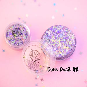 May include: Three clear plastic jars of glitter on a pink surface. The jars contain iridescent glitter in shades of purple, pink, and silver. One jar has a seashell graphic and the text "Facial Glitter, Hair Glitter, Body Glitter". The text "Diva Duck" is also visible.