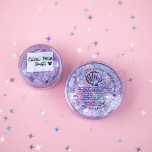 May include: Two clear circular jars filled with iridescent lilac and purple chunky glitter. One jar features a white label with the black text Lilac Pixie Dust and a mouse icon. The other jar lid displays a seashell icon and ingredient text.