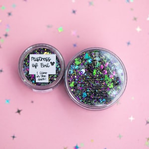 May include: Two clear circular containers filled with a mix of black, purple, and neon green glitter. The smaller container has a white label reading Mistress of Evil with a mouse icon and I Glow in the dark. The larger container features a shell icon.