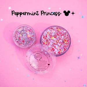 May include: Three clear circular containers filled with Peppermint Princess glitter in shades of pink, red, and white. The lids feature a seashell icon and text listing ingredients like beeswax, cocoa seed butter, and shea butter for hair and body.
