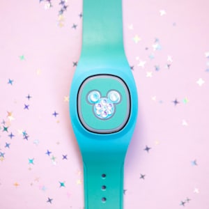 May include: A teal smart wristband with a Mickey Mouse design on the face. The band is a solid teal color with a light teal face. The Mickey Mouse design is made of small, iridescent gems. The background is a light pink with star-shaped confetti.