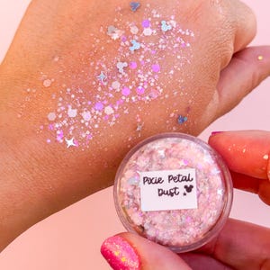 May include: A clear, round container of pink glitter labeled "Pixie Petal Dust" with a Mickey Mouse icon. The glitter is scattered on a hand, featuring various shapes and colors, including stars and Mickey Mouse silhouettes.
