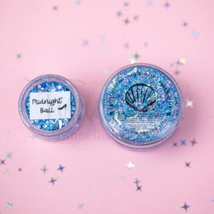 May include: Two clear circular containers filled with blue and iridescent cosmetic glitter. The smaller container has a white label reading Midnight Ball with a shoe icon. The larger container features a black seashell graphic and text.