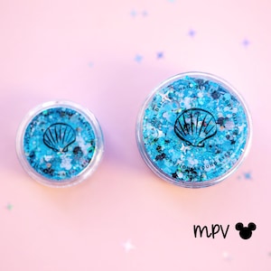 Puede incluir: Dos recipientes de plástico transparente llenos de purpurina azul. Cada recipiente tiene un gráfico de concha negra. El texto en los recipientes dice "FACIAL GLITTER HAIR GLITTER BODY GLITTER CAUTION AROUND EYES". El fondo es rosa.