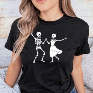 May include: Black t-shirt featuring a white graphic of two dancing skeletons. The skeletons are holding hands and are in a dancing pose. The shirt is made of a soft material and is suitable for casual wear.