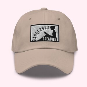 Puede incluir: Gorra de béisbol beige con visera curva. La gorra presenta un parche rectangular con las palabras "DANGEROUS CREATURE" y una silueta de una mujer leyendo.