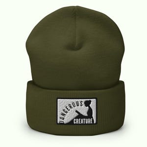 May include: Olive green beanie with a folded brim. A rectangular patch features the words "DANGEROUS CREATURE" around a silhouette of a person reading. The beanie is made of a soft, knitted material.