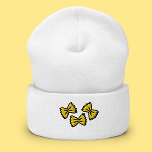 Pode incluir: Gorrinho branco com três formas de massa farfalle amarelas bordadas. O gorro é feito de um material macio e tricotado e tem uma bainha dobrada. As formas de massa são delineadas em preto.